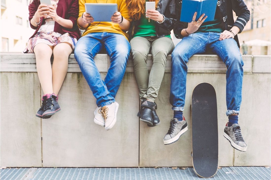 Four young people sitting on a wall with phones, a tablet and a book